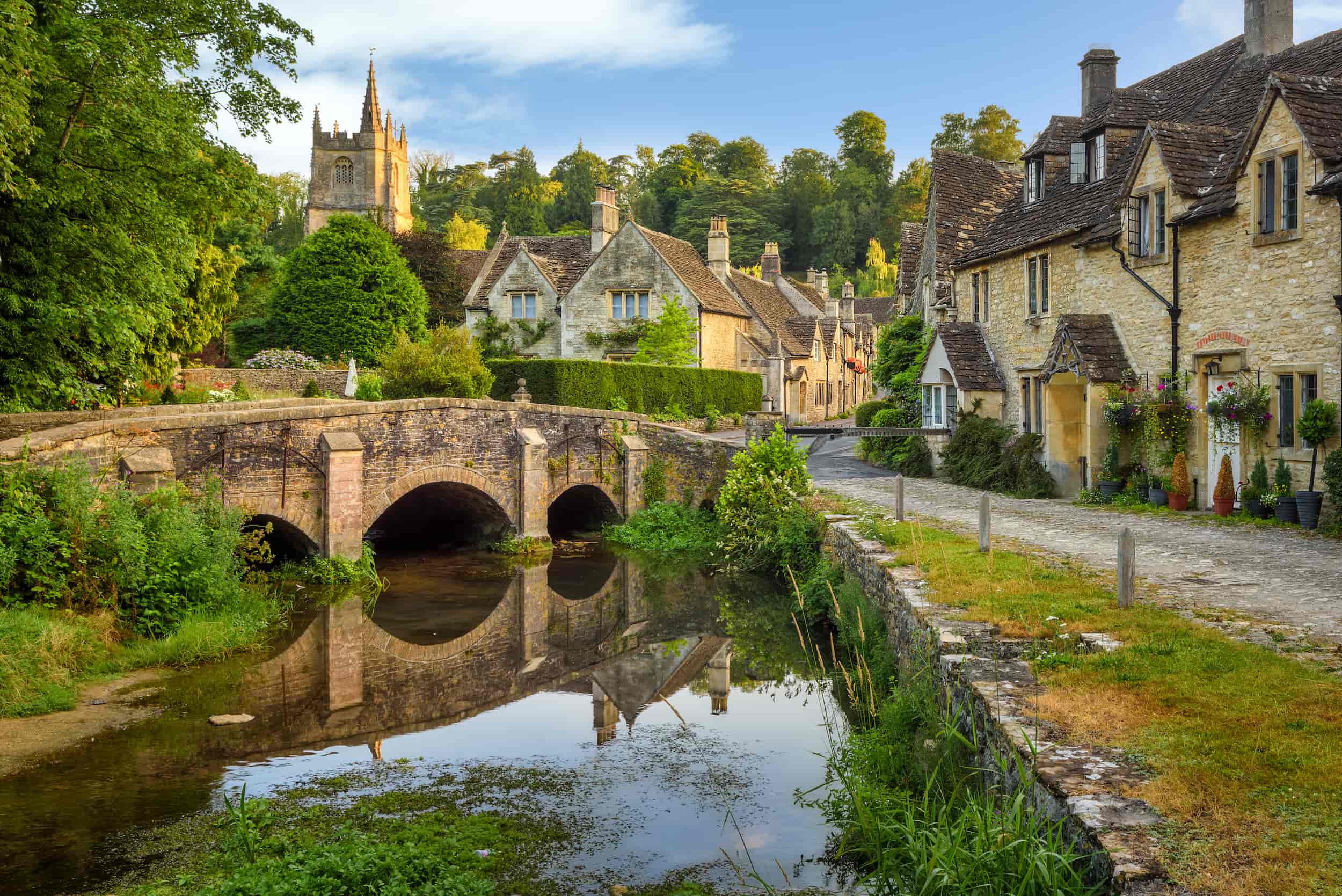 A stone bridge over a small river with historic stone cottages and lush greenery; a church with a tall spire rises in the background under a blue sky.