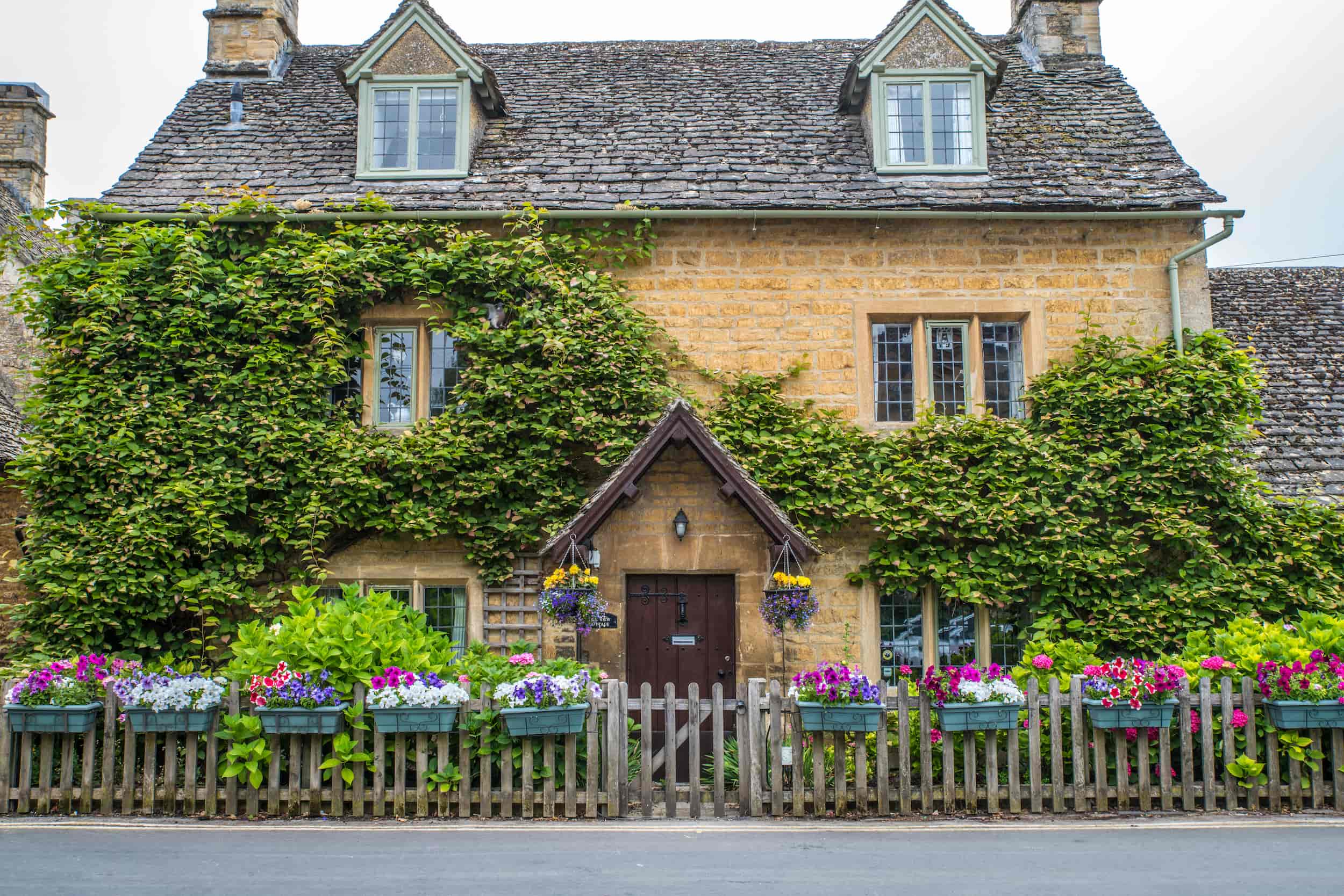 A charming stone cottage with ivy climbing its walls, dormer windows, and colorful flower boxes along a wooden fence and windowsills. A small peaked porch shelters the front door.