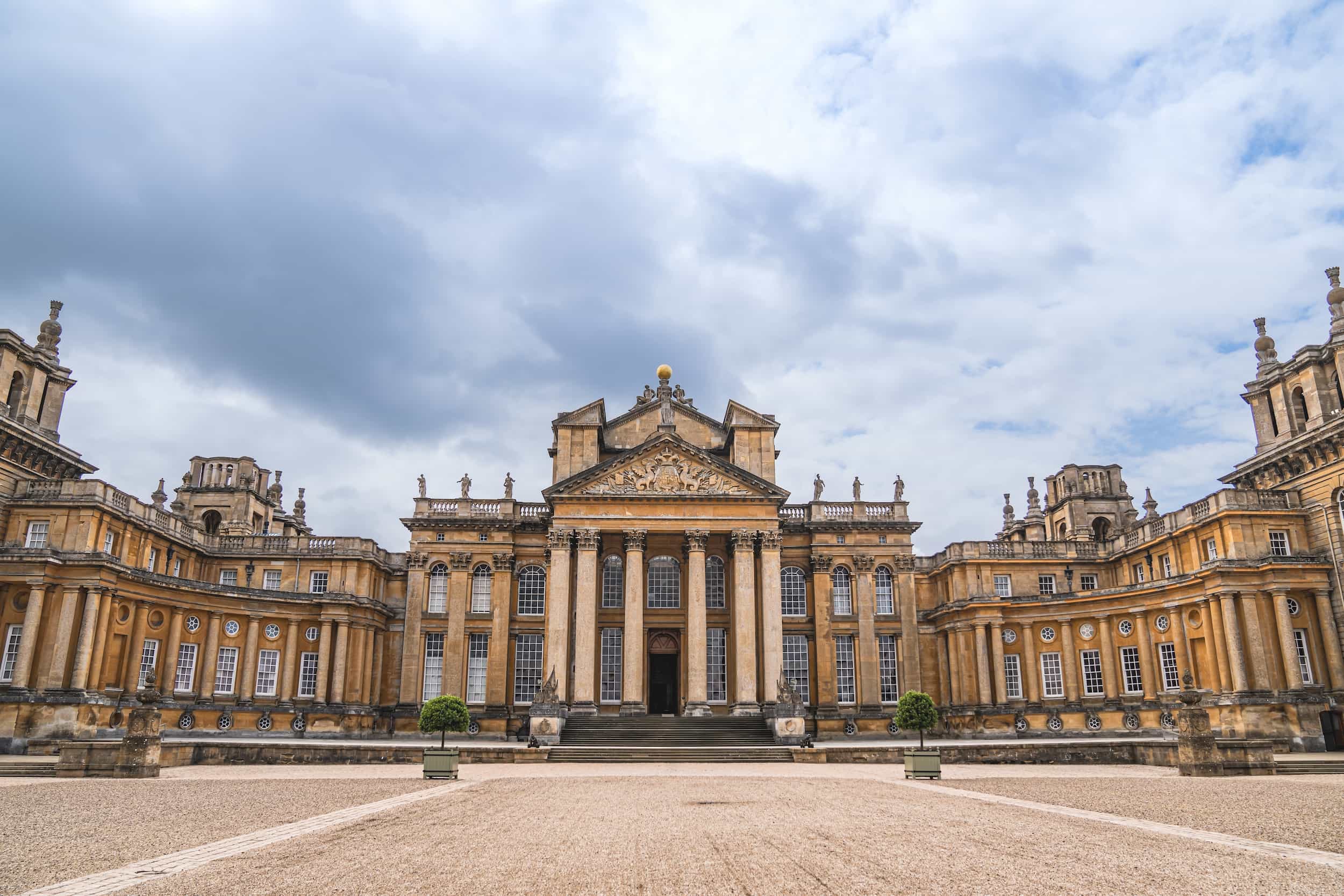 A grand historic palace with ornate columns, symmetrical wings, and detailed stonework, set under a cloudy sky. The building features statues on the roof and a central pediment above the main entrance.