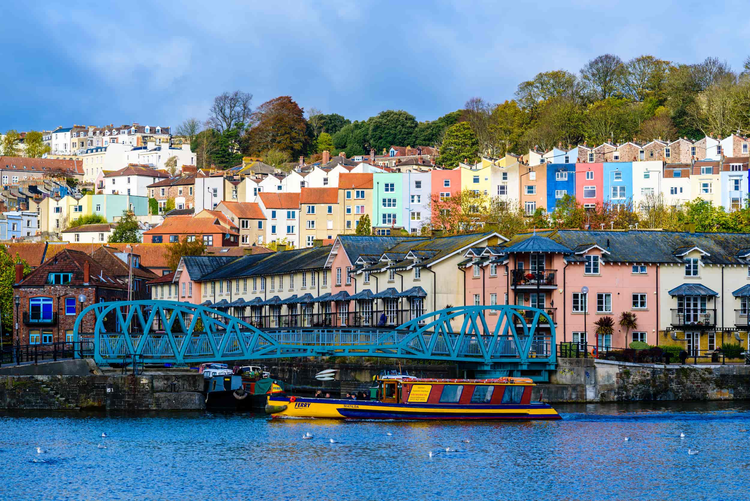 Colorful houses line a hillside above a blue metal bridge and waterfront buildings in Bristol, UK, with a yellow and purple boat floating on the river in the foreground.
