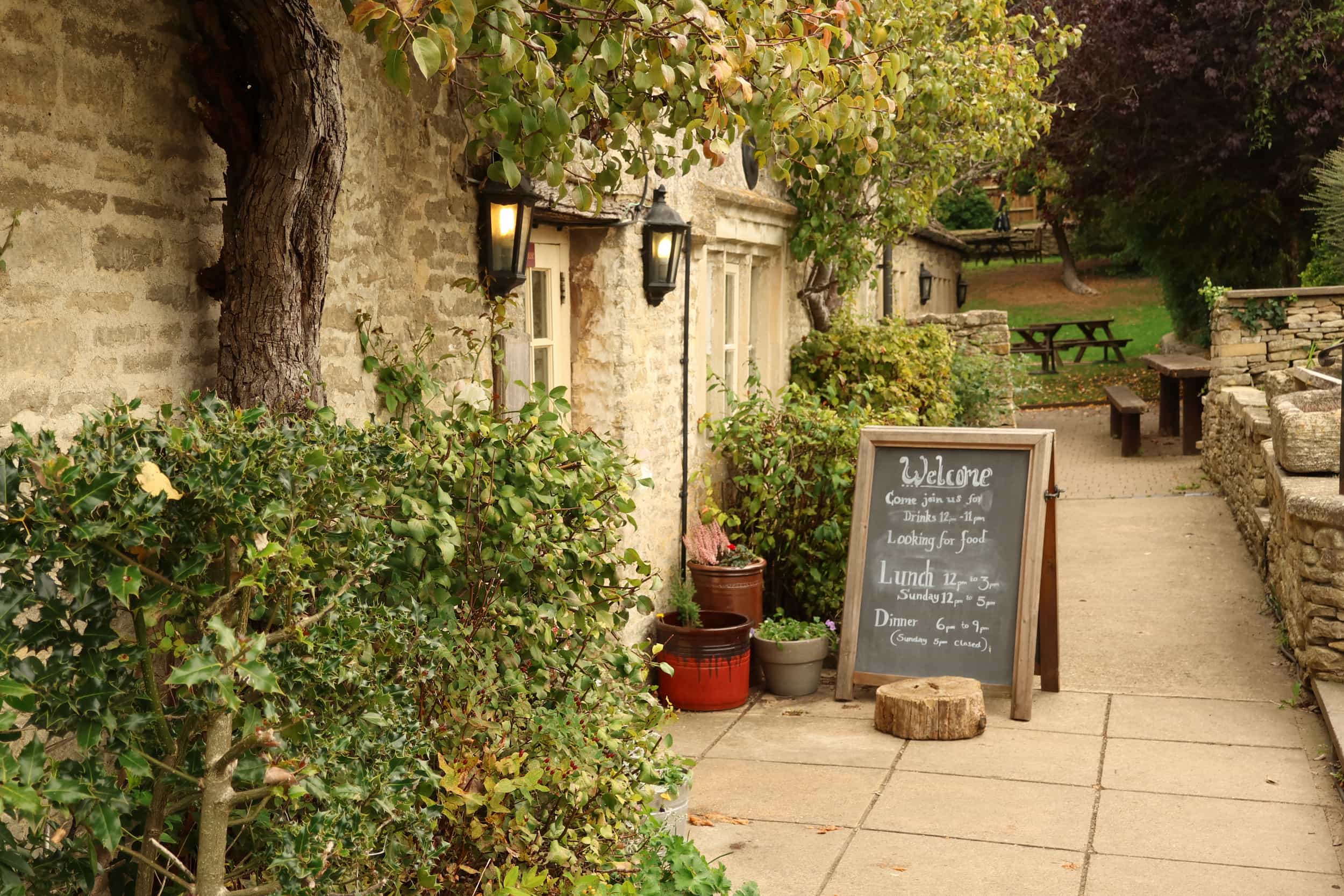 A rustic stone building with lanterns and greenery out front. A chalkboard menu welcomes guests and lists lunch specials. Wooden picnic tables are set up in the garden area further down the path.