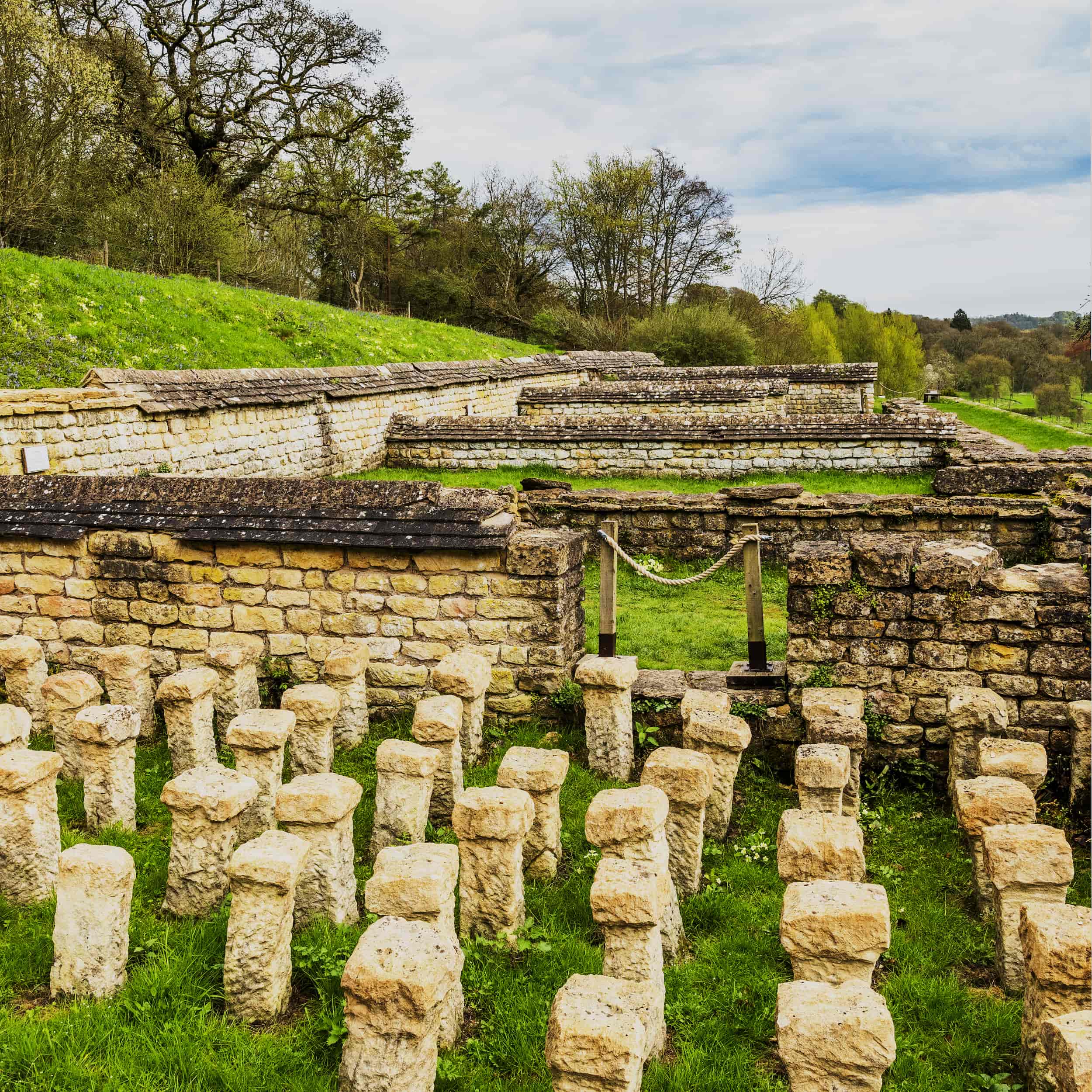 Ancient stone ruins with short, upright columns in the foreground, likely part of a Roman bathhouse or similar structure, surrounded by grassy hills and trees under a cloudy sky.