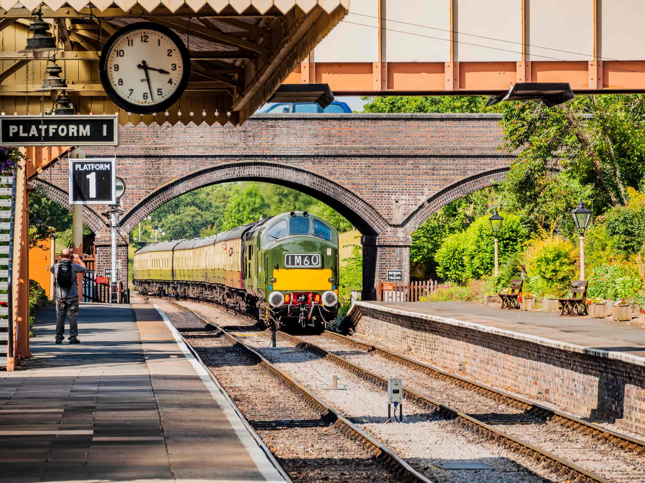 A vintage green train approaches Platform 1 at a quaint railway station, passing under a brick arch bridge. A person stands on the platform near benches and a clock showing 10:11. Trees and greenery surround the scene.