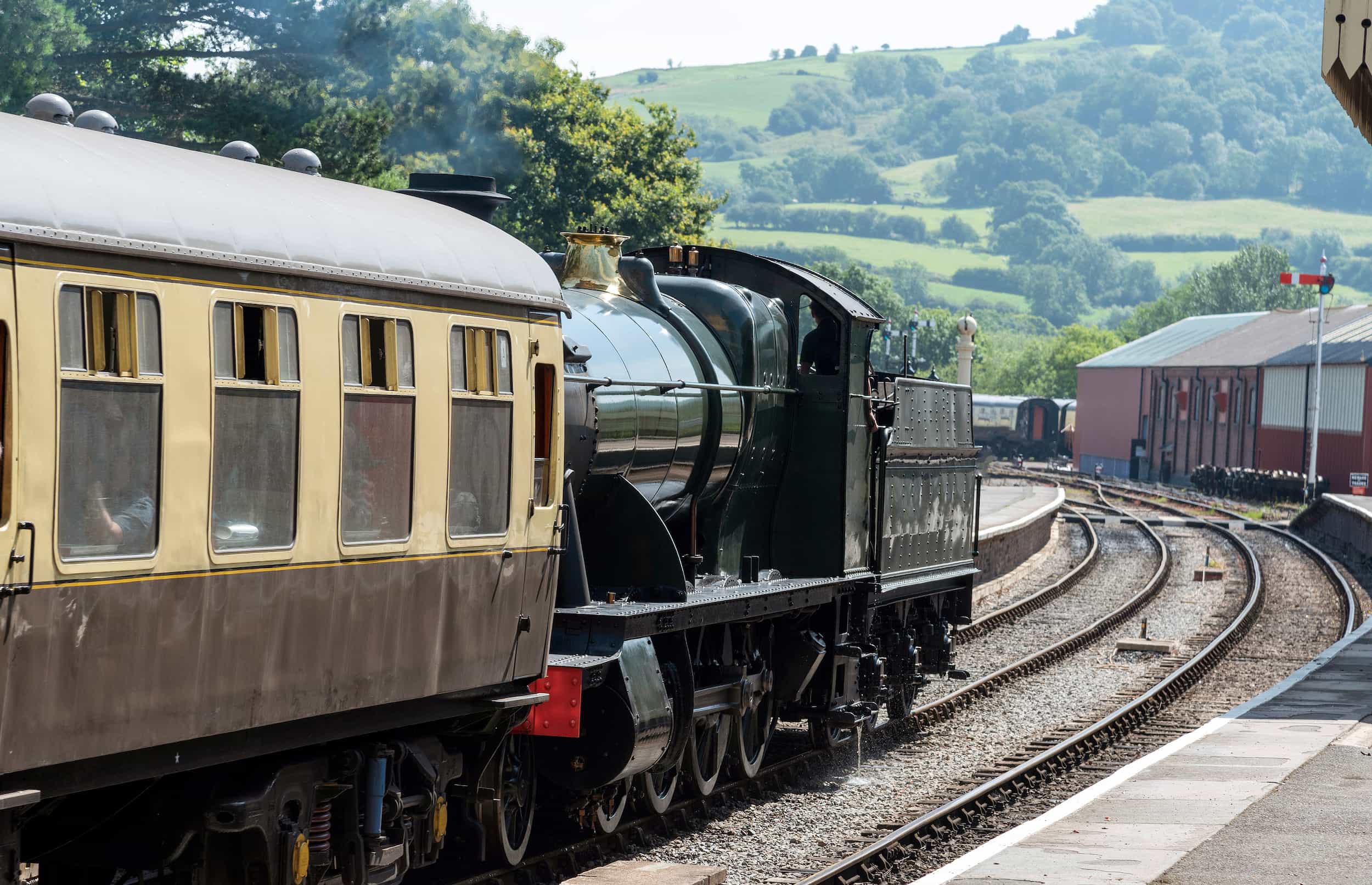 A vintage steam train with cream and brown carriages arrives at a rural station, with green hills and trees visible in the background under a sunny sky.