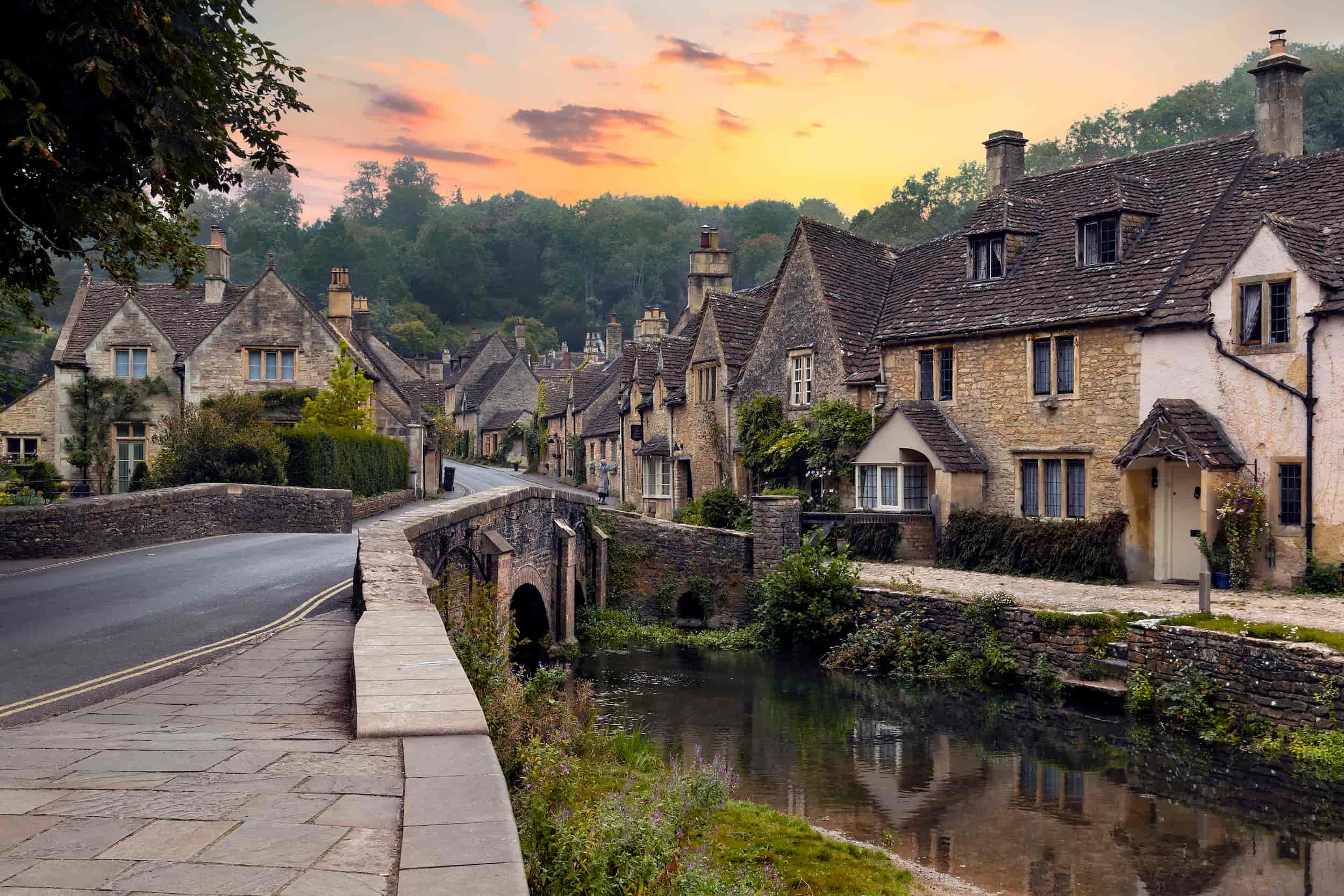 Charming stone cottages line a quiet village street beside a small river and stone bridge at sunset, surrounded by greenery and trees under a colorful sky.