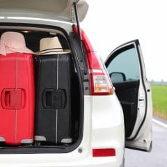 The back of a white car is open, showing several suitcases and hats packed inside. The car is parked on the side of a road next to a green field, with one rear door open.