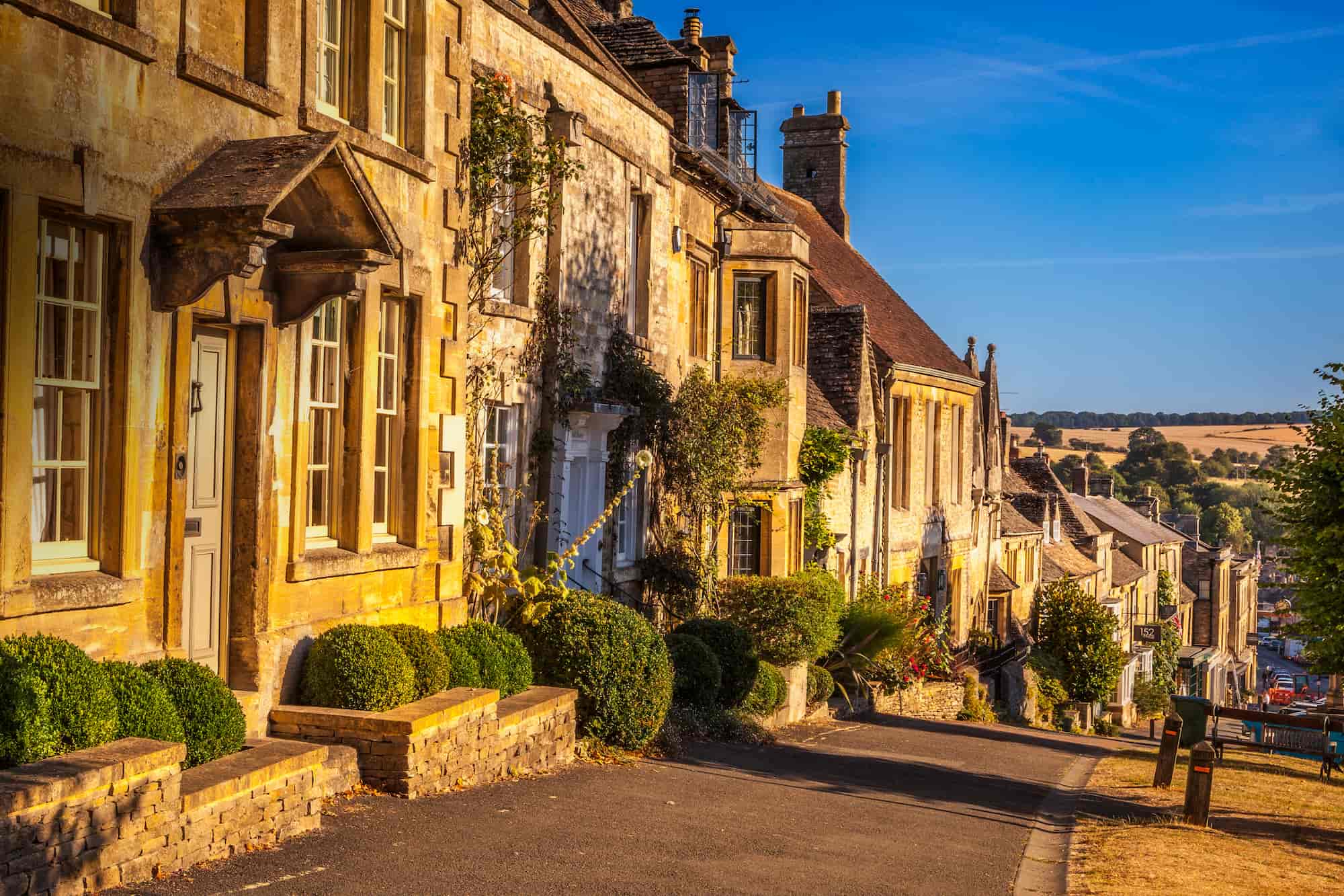 A sunlit street in a historic English village, lined with charming stone cottages, green bushes, and old-style windows, with rolling countryside visible in the background under a clear blue sky.