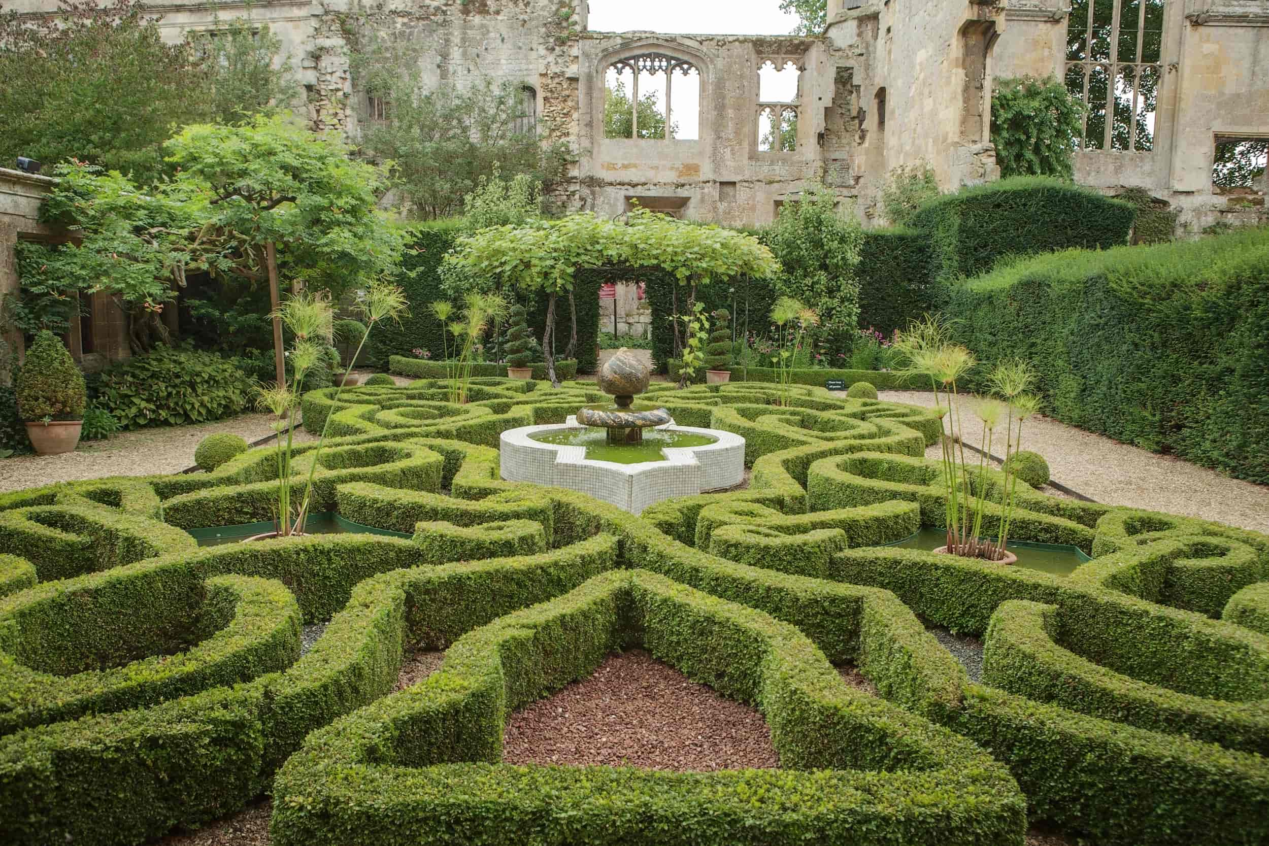 A formal garden with neatly trimmed, intricate boxwood hedges, a central fountain, gravel paths, and lush greenery, set against the backdrop of ancient, partially ruined stone walls.