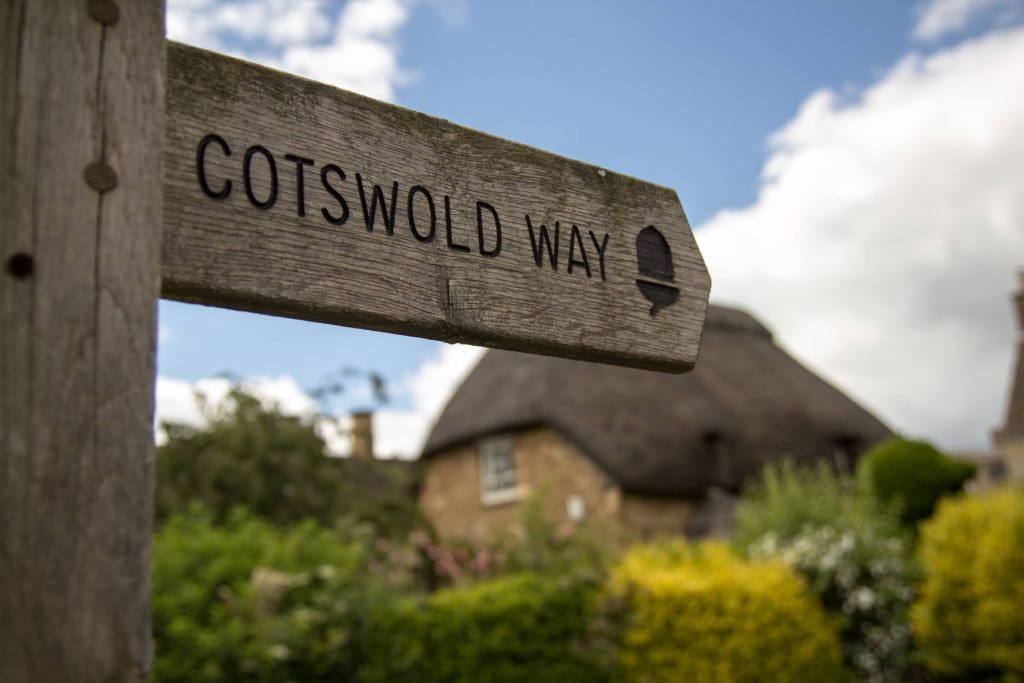 A wooden signpost reading "Cotswold Way" with an acorn symbol, set against a blurred background of a thatched-roof cottage, greenery, and a partly cloudy sky.