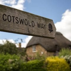 A wooden signpost reading "Cotswold Way" with an acorn symbol, set against a blurred background of a thatched-roof cottage, greenery, and a partly cloudy sky.