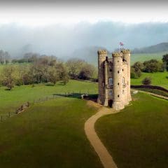 A small stone tower with three circular turrets stands on a grassy hilltop, surrounded by fields and trees. A path leads to the tower, and a British flag flies above. Misty woods are visible in the background.