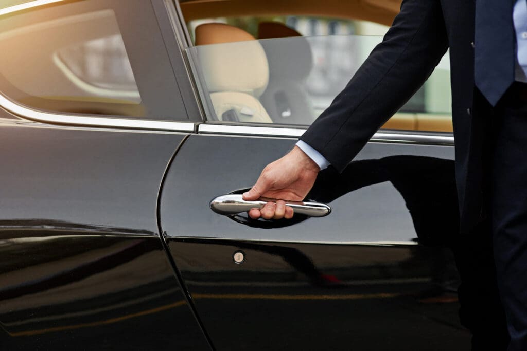 A person in a dark suit opens the door of a black luxury car. The car's interior and window are visible, and sunlight shines on the vehicle.