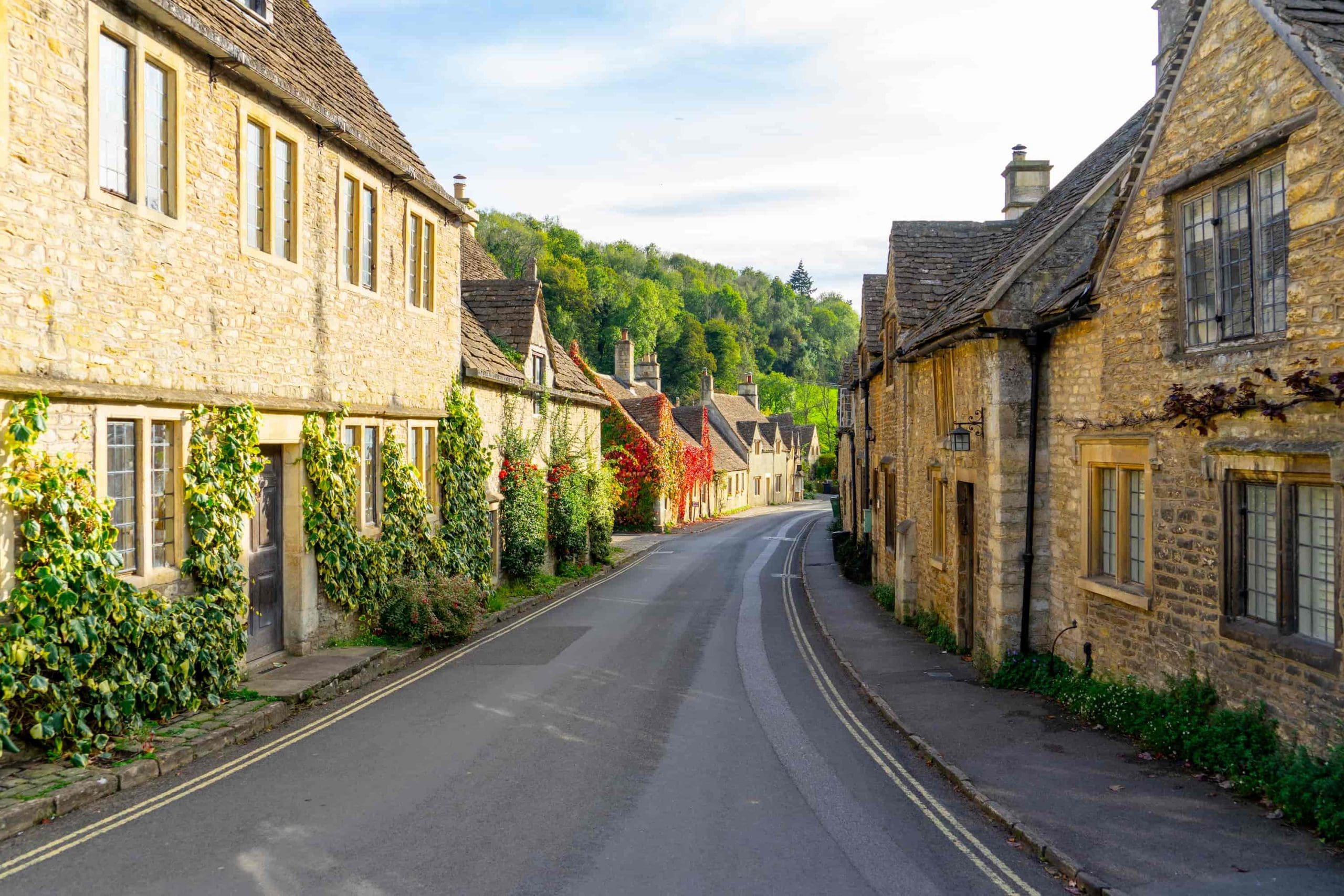 A quiet street lined with charming old stone houses covered in greenery, curving through a picturesque village with trees and hills in the background under a partly cloudy sky.