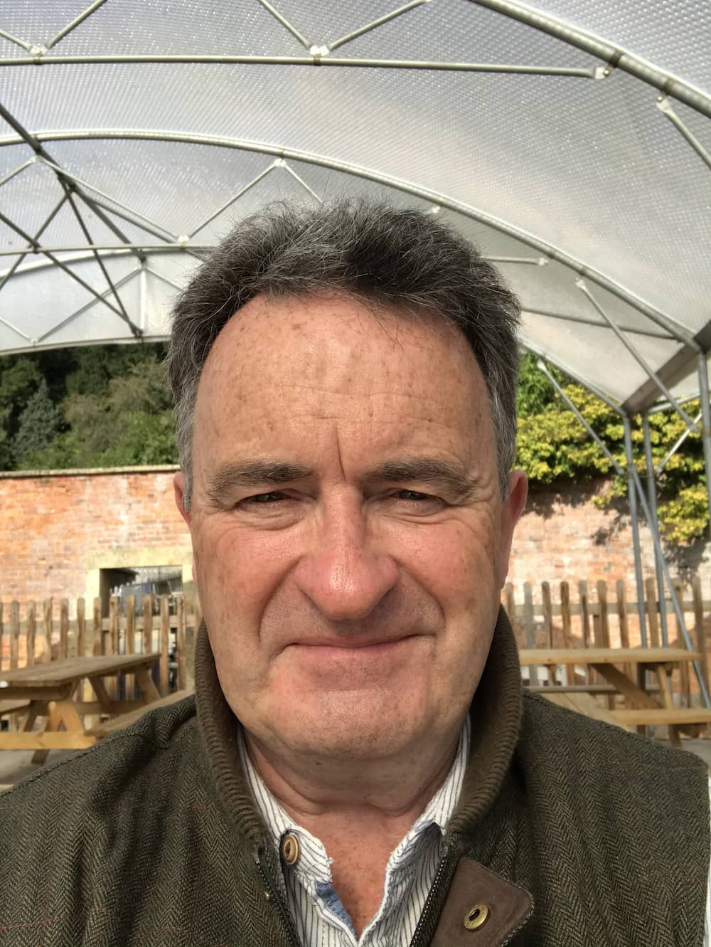 A middle-aged man with gray hair smiles outdoors under a clear canopy, with wooden picnic tables and a brick wall with greenery in the background.