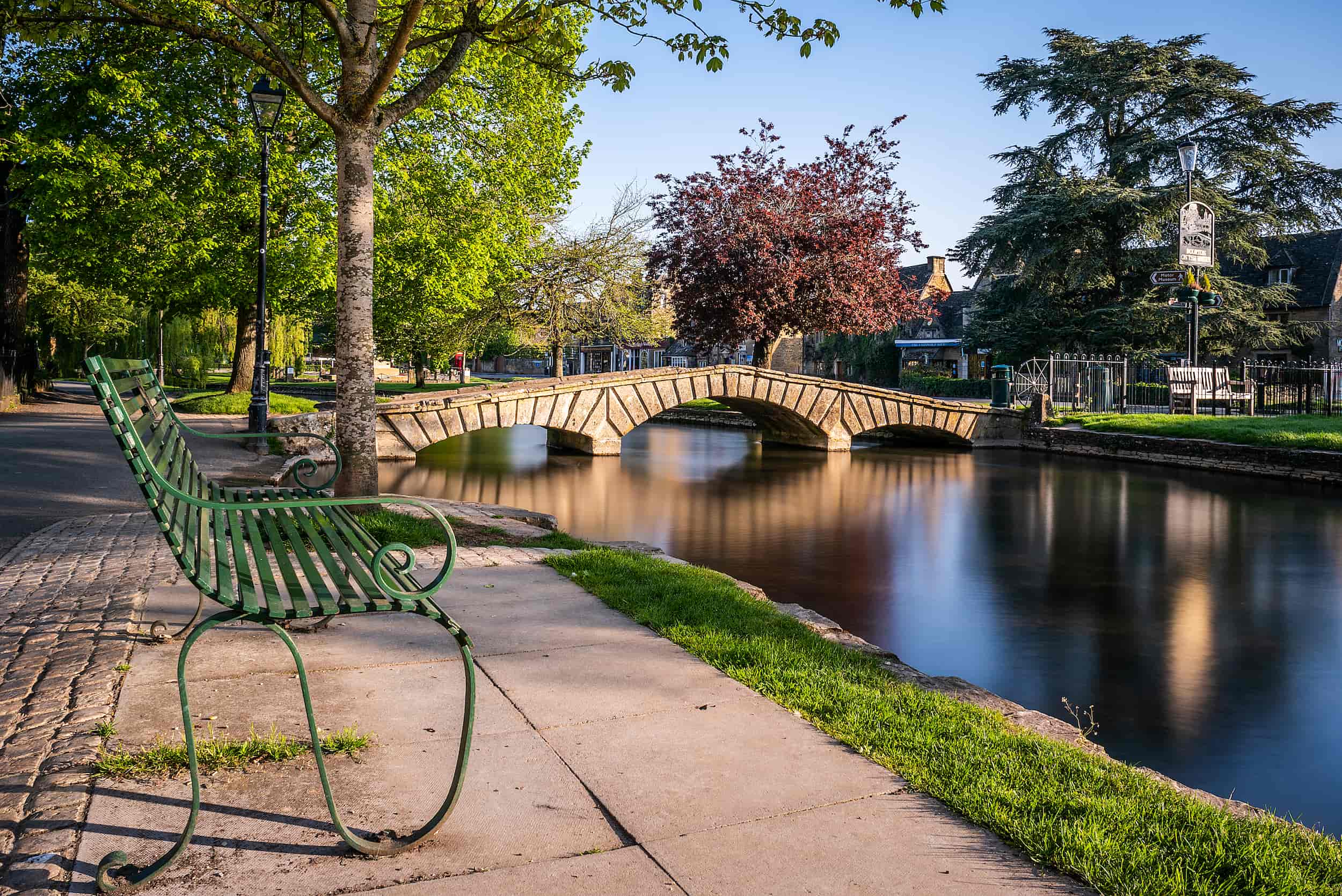 A green metal bench sits beside a calm river, with a stone footbridge crossing the water and lush trees lining the banks under a clear blue sky.