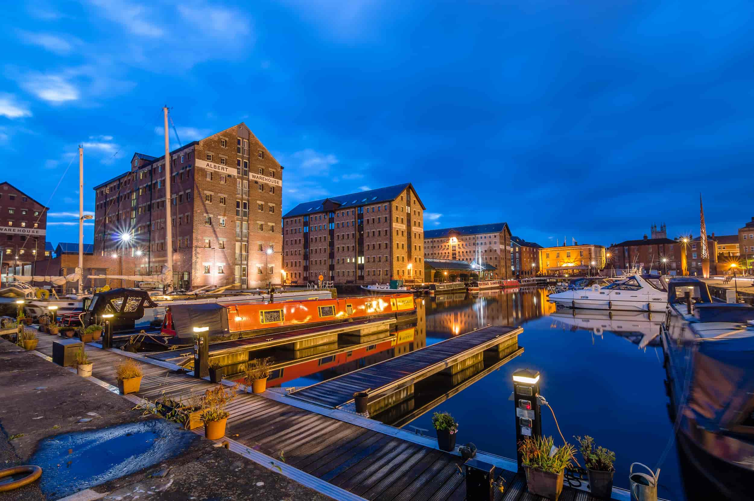 A waterfront scene at dusk with historic brick warehouses, lit-up boats and narrowboats moored along wooden docks, reflected in calm water under a deep blue sky.