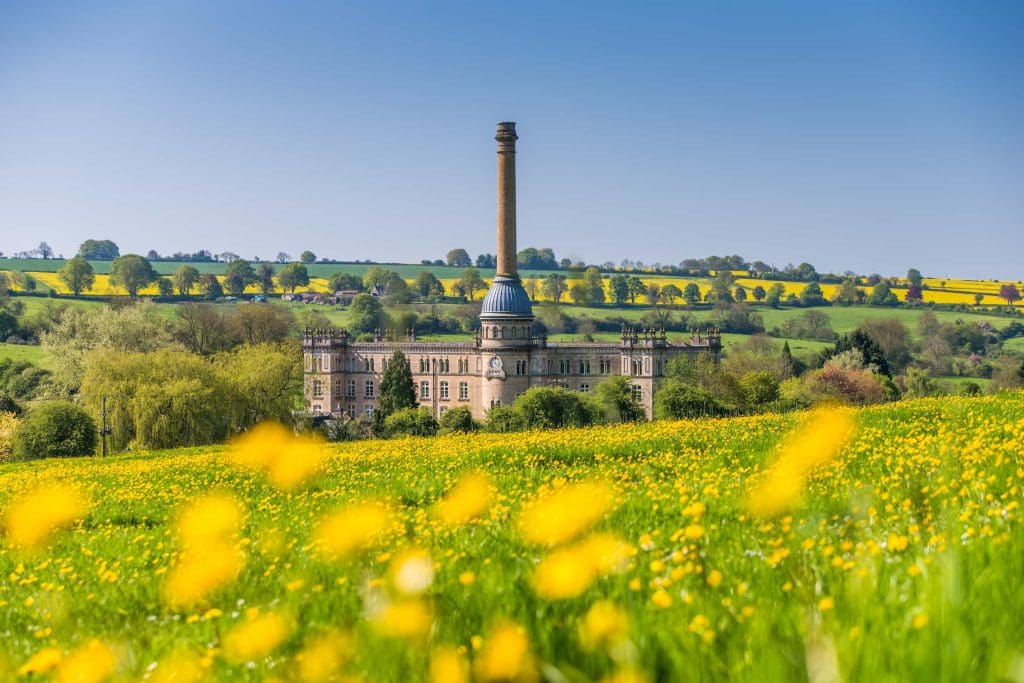 A historic mill building with a tall chimney stands amid green fields and yellow wildflowers under a clear blue sky, with rolling hills and trees in the background.