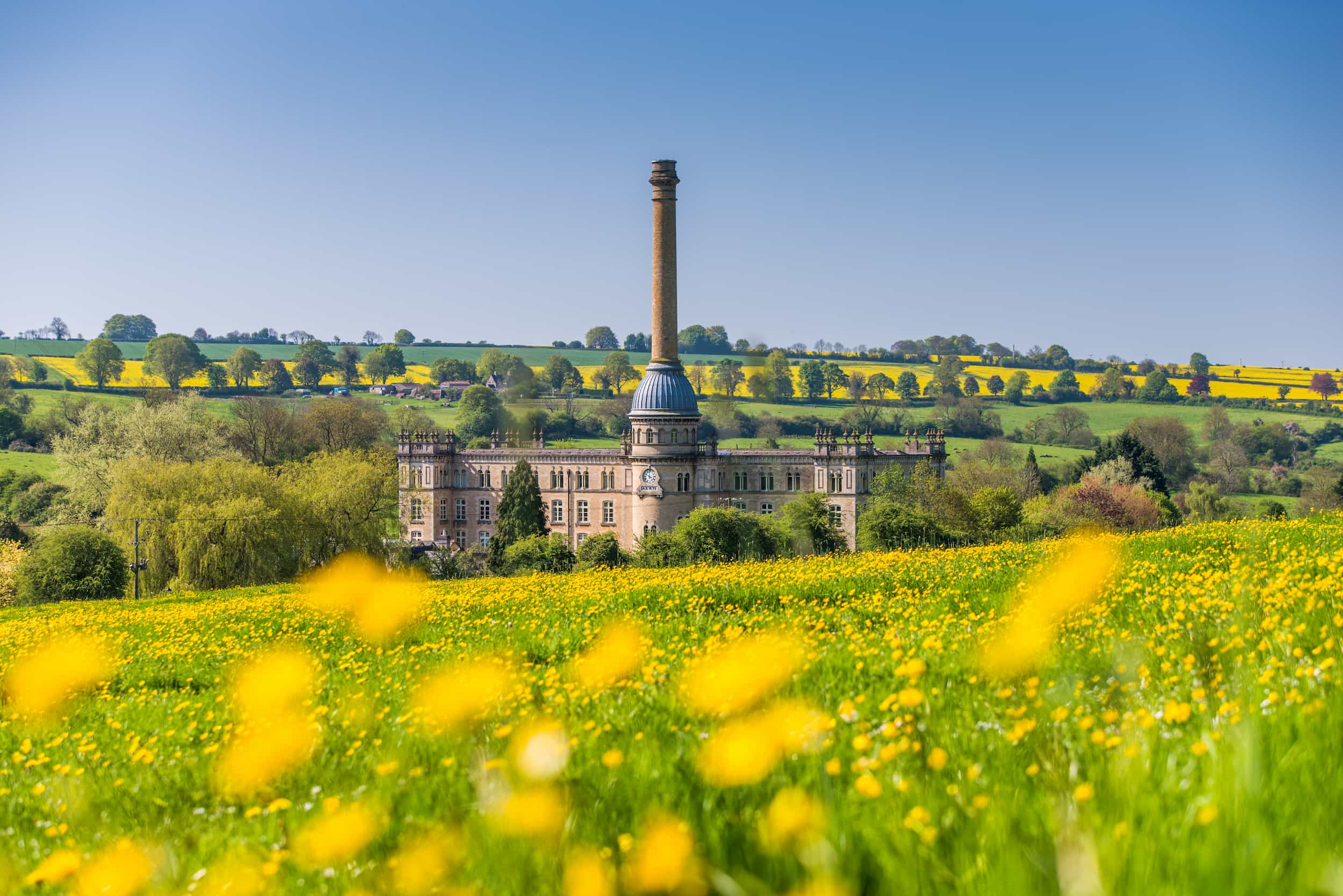 A historic mill building with a tall chimney stands amid green fields and yellow wildflowers under a clear blue sky, with rolling hills and trees in the background.