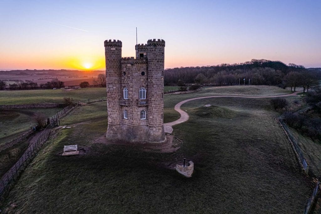 Aerial view of a small, historic stone tower with two round turrets, set on grassy open land at sunset, with a path leading to the tower and the sun low on the horizon.
