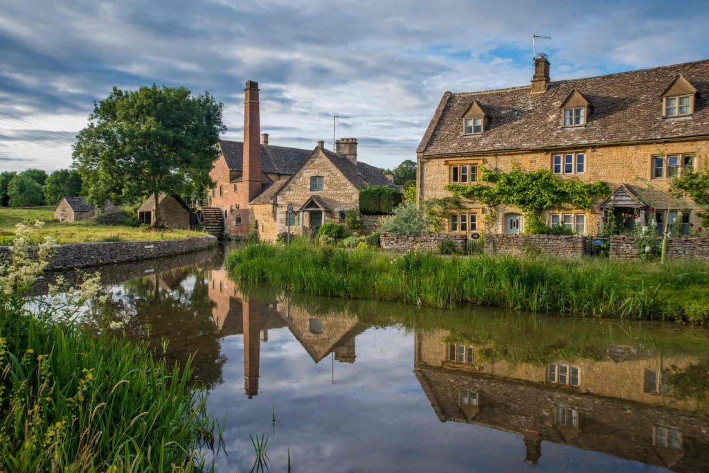 A tranquil village scene with stone cottages by a calm river, lush greenery, and a tree, all reflected in the water under a partly cloudy sky.