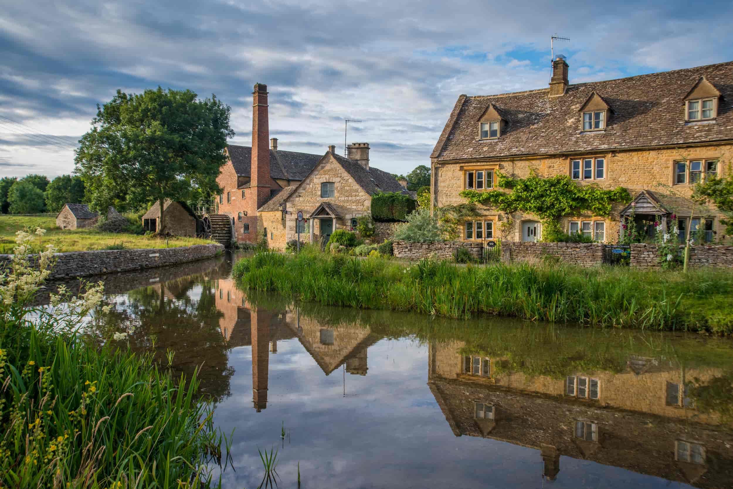 A tranquil village scene with stone cottages by a calm river, lush greenery, and a tree, all reflected in the water under a partly cloudy sky.