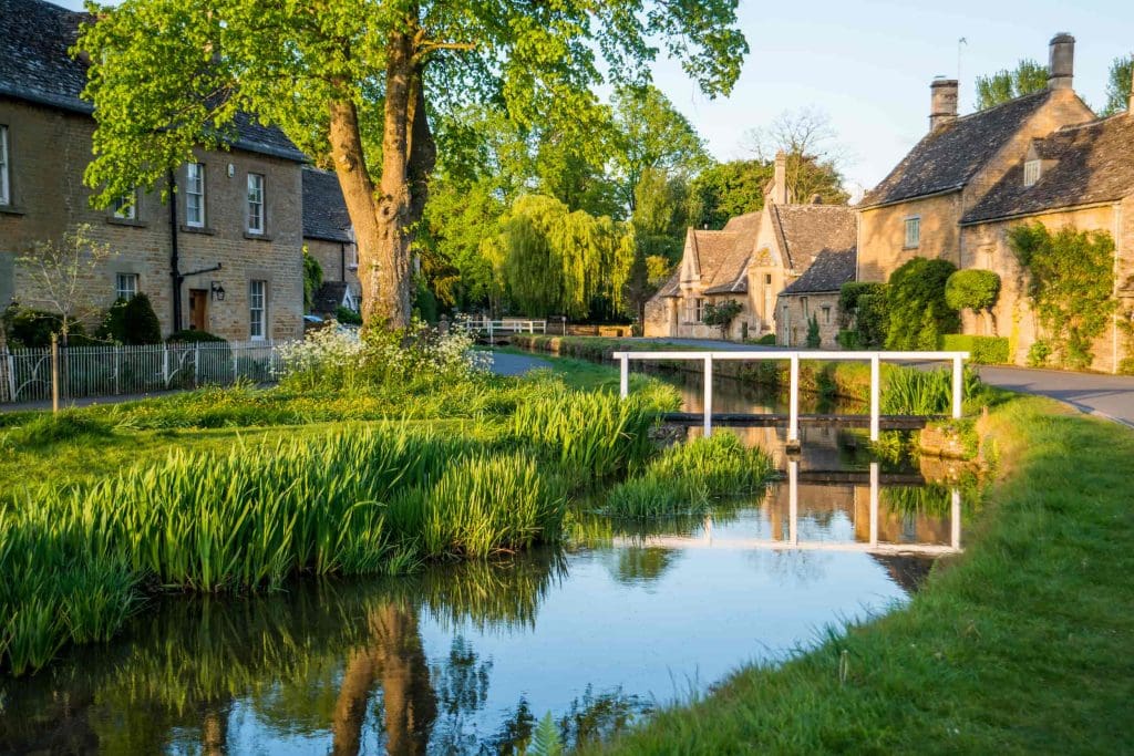 A picturesque village scene with stone cottages, a small arched white bridge over a calm stream, lush green grass, and tall trees under bright sunlight.