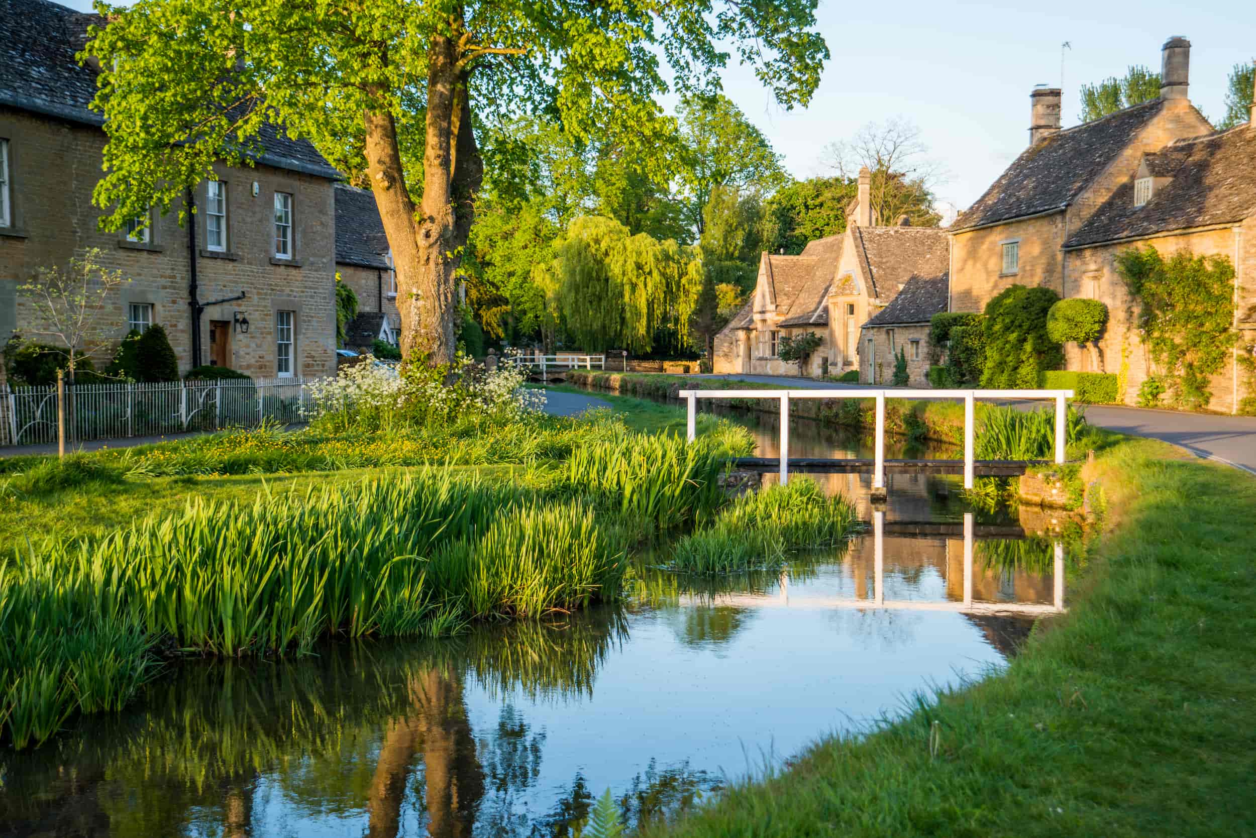 A picturesque village scene with stone cottages, a small arched white bridge over a calm stream, lush green grass, and tall trees under bright sunlight.