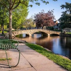 A green metal bench sits beside a calm river, with a stone footbridge crossing the water. Trees with green and red leaves line the riverbank under a clear blue sky.