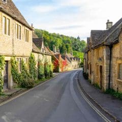 A quiet, narrow street lined with charming stone cottages, ivy climbing the walls, and green hills in the background under a partly cloudy sky.