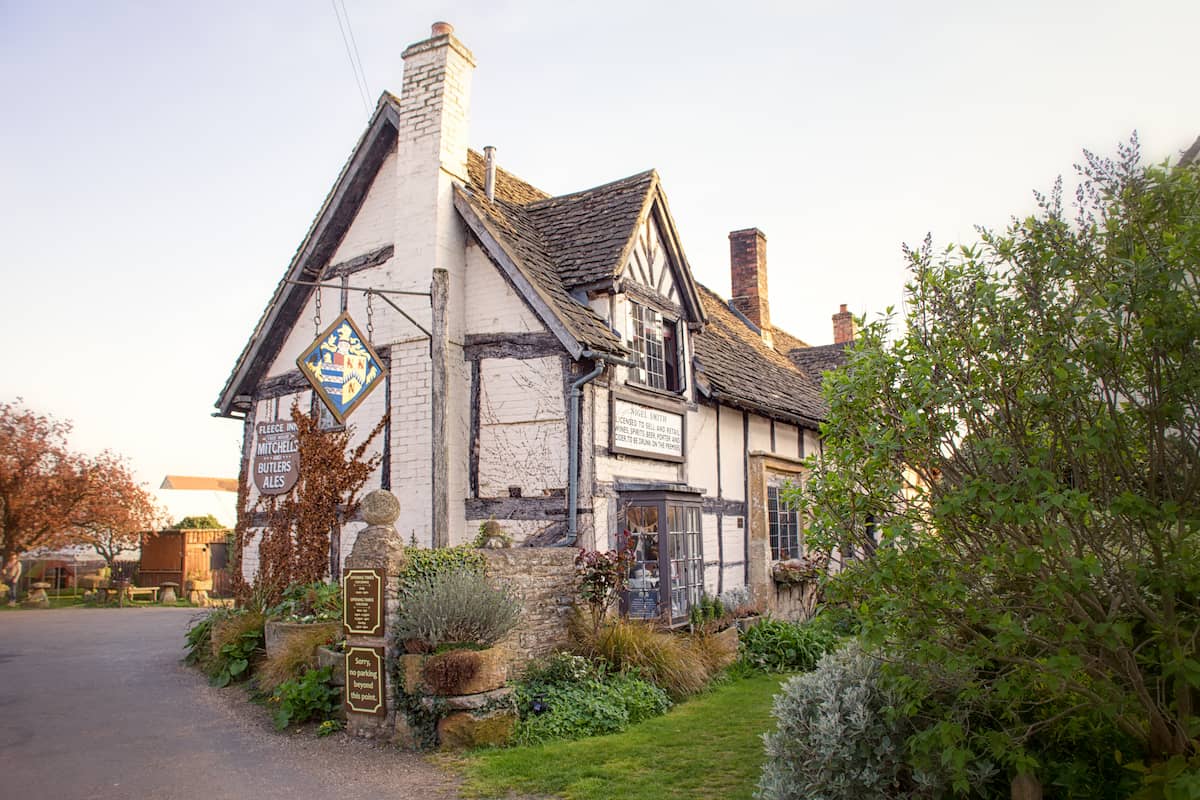 A charming old English pub with timber framing, a stone chimney, and slate roof, surrounded by lush greenery and signage by the entrance on a sunny day.